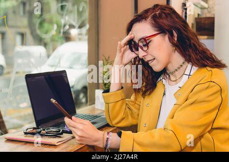Rothaarige junge Frau mit Brille und gelbem Blazer freiberuflich mit ihrem Laptop in einem Café arbeiten und empört auf das Telefon schauen. Stockfoto