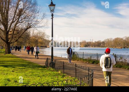 Menschen, die an einem altmodischen Laternenpfosten auf dem Fußweg am Serpentine Lake im Hyde Park, London, an einem hellen sonnigen Wintertag spazieren gehen. Stockfoto