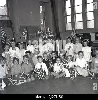 1965, historische, aufgeregt Gruppe von Grundschulkindern, die in einer Schulhalle mit Union Jack Flaggen sitzen, Fife, Schottland, Großbritannien. Stockfoto