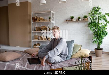Ein junger, gutaussehender Mann sitzt auf einem Bett neben einem Laptop. Schaut auf die Kamera und lächelt Stockfoto