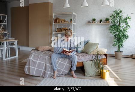 Ein hübscher junger Mann sitzt am Rand des Bettes und liest ein Buch, während er sich in seinem Schlafzimmer entspannt. Stockfoto