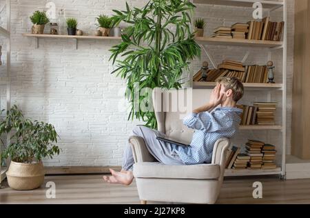 Müde von der Arbeit an einem Laptop sitzt ein Mann auf einem Stuhl, der sein Gesicht mit den Händen bedeckt Stockfoto