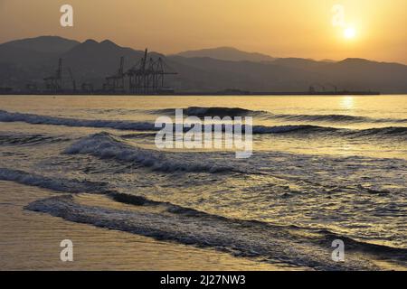 Wellen nähern sich der Küste von Malaga in der Morgendämmerung mit Industriehafen im Hintergrund, Costa del Sol Andalusien Südspanien. Stockfoto