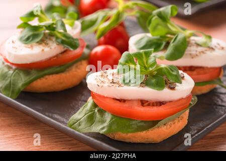 Nahaufnahme der klassischen italienischen Vorspeise - Caprese-Salat mit geschnittenen Tomaten, Mozzarella, Basilikum und gekrönt mit Mais-Salat. Bruschettas auf schwarzem pla Stockfoto