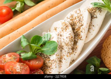 Nahaufnahme des traditionellen italienischen Caprese-Salats mit Babytomaten und Mozzarella in Scheiben, Basilikum, Olivenöl, Maissalat, Bruschettas und Grissini Stockfoto