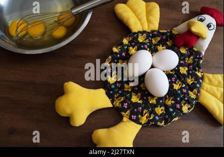 Drei Eier auf einem Hühnchen aus Stoff und vier Eigelb mit Weiß in einer Metallschüssel mit einem Schneebesen darin Stockfoto