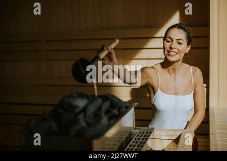 Attraktive junge Frau, die in der Sauna Wasser auf heißen Stein gießt Stockfoto