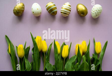 Ostereier aus Gold, weiße Farbe mit Muster und gelbe Tulpen auf violettem Hintergrund. Speicherplatz kopieren. Stockfoto