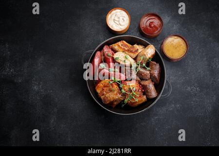 Gebratene Fleischplatte in einer Pfanne und Saucen, Würstchen, Rippchen, Hähnchenwurst, Schweinewurst, Auf schwarzem Hintergrund Stockfoto