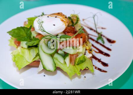 Toast mit Lachs und pochiertem Ei. Salat, Gurke, Basilikum. Ein gesundes Frühstückssandwich. Microgreen und Flachs- und Sesamsamen. Gesundes Essen auf einem weißen Teller. Fisch mit Gemüse auf Brot. Stockfoto