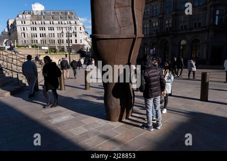 Menschen interagieren mit der Iron:man-Statue des britischen Bildhauers Antony Gormley am 14.. März 2022 auf dem Victoria Square in Birmingham, Großbritannien. Die Statue ist 6 Meter hoch, einschließlich der Füße, die unter dem Bürgersteig begraben sind, und wiegt 6 Tonnen. Die Statue lehnt sich um 7,5 Grad nach hinten und um 5 Grad nach links. Es wird vom Bildhauer gesagt, die traditionellen Fähigkeiten von Birmingham und dem Schwarzen Land zu repräsentieren, die während der industriellen Revolution praktiziert wurden. Die Statue wurde im September 2017 gelagert, um die Gleise für die U-Bahn-Erweiterung der West Midlands zum Centenary Square zu legen, und Stockfoto