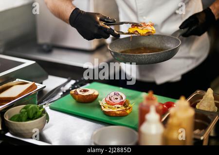 Ansicht der männlichen Hände in Handschuhen mit saftigem gegrilltem Kitt mit Käse und Speck, mit Zange und Pfanne. Zubereitung von leckeren Cheeseburger mit Saucen auf dem Hintergrund. Konzept von Fast Food. Stockfoto