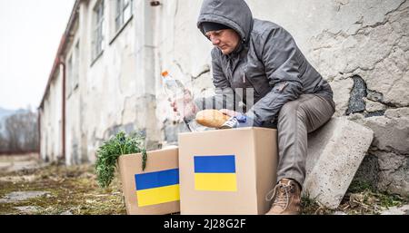 Ein älterer Mann in warmen Kleidern sitzt am Rande eines entgleisten Gebäudes und nimmt Brot und Wasser aus dem humanitären Hilfspaket für die Ukraine. Stockfoto