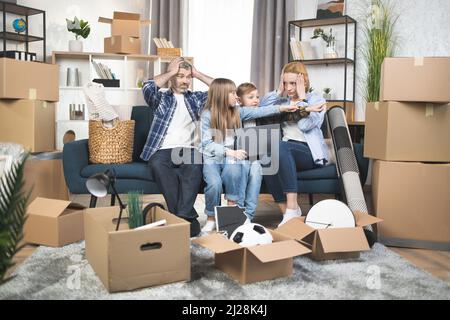 Zwei Kinder helfen Eltern, nach einem Umzug Kisten zu berechnen. Schockierte Eltern, die auf der Couch saßen und den Kopf mit den Händen berührten, um sich auf den Tag vorzubereiten. Vergessene Schachtel, Dinge verlieren Stockfoto