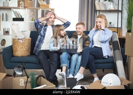 Glücklich motivierte Kinder helfen Eltern, Kisten für den Umzug zu packen. Besorgte Eltern sitzen auf der Couch in Erwartung großer Veränderungen und bereiten sich auf den Tag vor. Besitzer einer neuen modernen Wohnung. Stockfoto