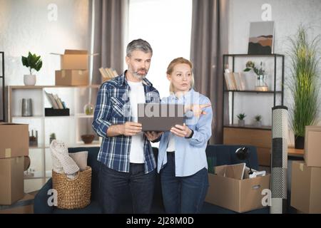Kaukasisches reifes Paar, das zwischen Kartons und verschiedenen Dingen steht und einen Laptop verwendet, das Design sucht und Online-Shopping macht. Familie, die beim Entfernen in eine neue Wohnung Pause einnahm. Stockfoto