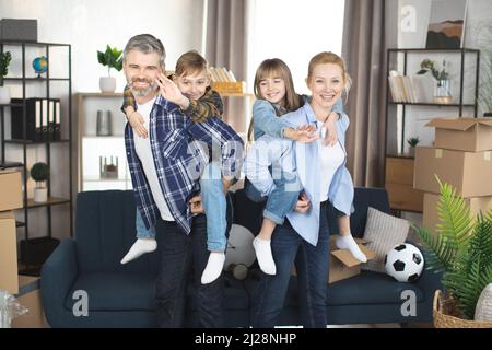 Glückliche Eltern Huckepack Sohn und Tochter zu Hause. Lächelnder Vater und Mutter halten ihre Kinder auf dem Rücken verbringen Zeit zusammen, Familie Spaß haben Umzug Umzug Tag Stockfoto