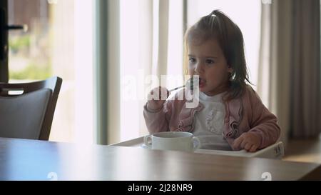 Nahaufnahme von jungen Baby in weiß Fütterung Hochstuhl, Kind versucht, sich selbst zu essen, glückliches Kind mit Essen verschmutztes Gesicht, kleines Mädchen isst Brei mit Stockfoto