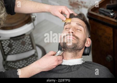 Junger Hipster kaukasischer Mann während der Bartpflege in einem modernen Friseurladen. Haarstyling für Männer. Schöner Mann immer neue Frisur mit elektrischen Trimmer. Hochwertige Fotografie Stockfoto