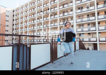 Tapferes, glückliches kaukasisches blondes Teenager-Mädchen in den Jahren 90s, das im Freien tanzt und ihren altmodischen Kassettenspieler in der Hand hält. Wohnblock im Hintergrund. Hochwertige Fotos Stockfoto