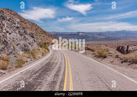 Nelson, Nevada, USA - 21. Mai 2011: Graue Asphaltroute 165 führt unter blauer Wolkenlandschaft zum blauen Wasser des Colorado River, während sie durch trockene Wüste führt Stockfoto