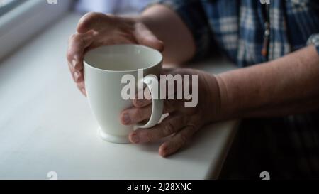 Senior hält eine Tasse Kaffee, während er am Fenster steht Stockfoto