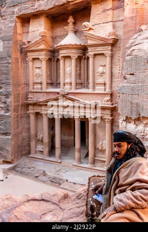 Ein lokaler Beduinenmensch in typischer Kleidung mit dem Äußeren des Schatzhauses ‘Al-Khazneh’ im Hintergrund, Petra, Jordanien, Asien. Stockfoto