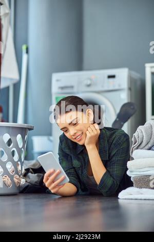 Sie überprüft ihre Nachrichten, während sie wartet. Aufnahme einer attraktiven jungen Frau, die ihr Telefon benutzt, während sie auf dem Boden liegt und auf die nächste Ladung Wäsche wartet Stockfoto