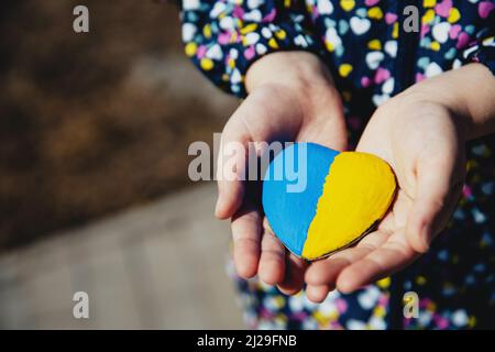 Kleines Kind halten herzförmigen Stein mit Ukraine blau-gelbe Nationalflagge oder Hymne. Mädchen lieben Heimat. Kinderstand für Ukrainer. Stoppt Krieg, Aggression, Invasion. Die Menschen verhals den Krieg. Stockfoto