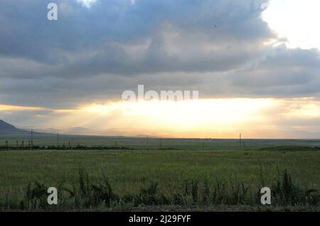 Die hellen Strahlen der untergehenden Sonne scheinen durch eine riesige Wolke im Abendtal in der hügeligen Steppe. Chakassien, Sibirien, Russland. Stockfoto