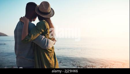 Ihre Liebe ist alles, was zählt. Rückansicht eines liebevollen jungen Paares, das sich am Strand bei Sonnenuntergang umarmt. Stockfoto