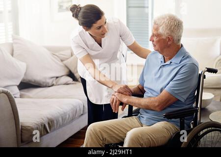 Ihm den ganzen Tag über zu helfen. Aufnahme eines Betreuers, der einem älteren Mann im Rollstuhl zu Hause hilft. Stockfoto