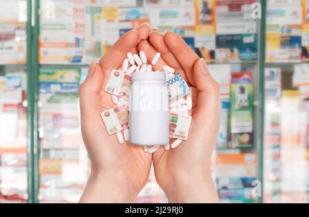 Nahaufnahme der Handflächen einer Frau mit einem weißen Glas mit Pillen und kleinen Rubeln. Im Hintergrund verschwommene Apothekenfenster. Das Konzept von Inflat Stockfoto