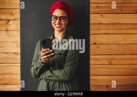 Geschäftsfrau lächelt in die Kamera, während sie ein Smartphone hält. Fröhliche muslimische Geschäftsfrau liest eine SMS in einem Büro. Glückliche weibliche Vorspeise Stockfoto