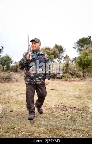 Durchgehender männlicher Jäger in Camouflage-Oberbekleidung und Mütze, der eine Waffe auf der Schulter trägt Stockfoto