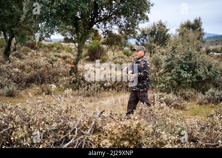 Männlicher Jäger in Camouflage-Oberbekleidung und Mütze, der eine Waffe trägt, die auf dem Feld läuft Stockfoto