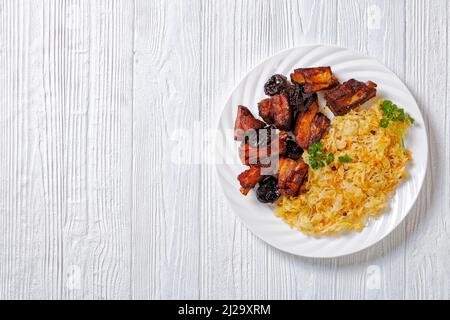 Geschmortes Sauerkraut mit Schweinefleisch und Pflaumen auf weißem Teller auf Holztisch, horizontale Ansicht von oben, flaches Lay, Freiraum Stockfoto