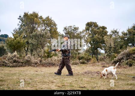 Männlicher Jäger in Tarnbekleidung und Mütze mit einer Waffe, der mit einem weißen Hund auf der Suche nach Beute auf dem Feld herumläuft Stockfoto