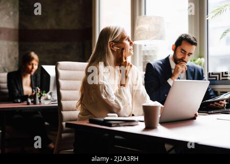 Eine reife Geschäftsfrau, die in-Ear-Kopfhörer am Laptop trägt, von einem Geschäftsmann am Schreibtisch im Büro Stockfoto