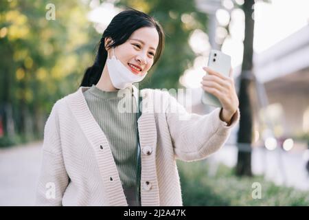 Asiatische Frau zieht ihre Gesichtsmaske aus, um ihr Telefon zu benutzen Stockfoto