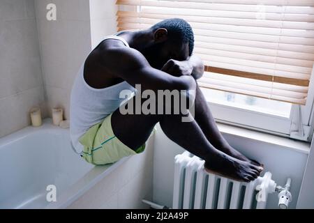 Die ganze Länge des einsamen Mannes, der unter Depressionen leidet, sitzt auf der Badewanne durch Jalousien im Badezimmer zu Hause Stockfoto