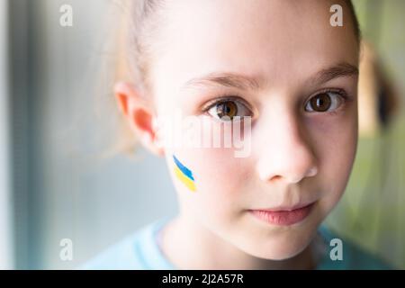 Gesicht eines verängstigten Mädchens, ein bemaltes Herz auf der Wange in gelb-blauen Farben der ukrainischen Flagge. Russlands Invasion der Ukraine, eine Bitte um Hilfe Stockfoto