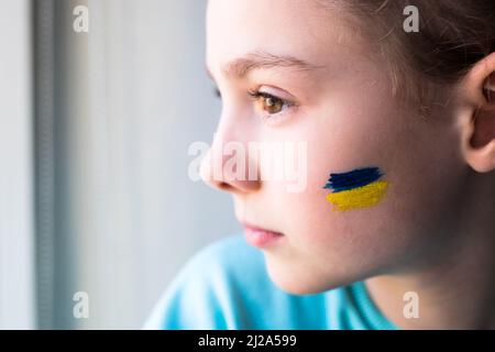 Gesicht eines verängstigten Mädchens, ein bemaltes Herz auf der Wange in gelb-blauen Farben der ukrainischen Flagge. Russlands Invasion der Ukraine, eine Bitte um Hilfe Stockfoto