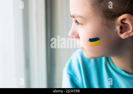 Gesicht eines verängstigten Mädchens, ein bemaltes Herz auf der Wange in gelb-blauen Farben der ukrainischen Flagge. Russlands Invasion der Ukraine, eine Bitte um Hilfe Stockfoto