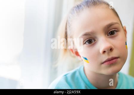 Gesicht eines verängstigten Mädchens, ein bemaltes Herz auf der Wange in gelb-blauen Farben der ukrainischen Flagge. Russlands Invasion der Ukraine, eine Bitte um Hilfe Stockfoto