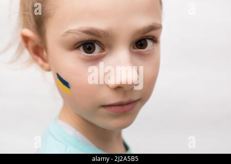 Gesicht eines verängstigten Mädchens, ein bemaltes Herz auf der Wange in gelb-blauen Farben der ukrainischen Flagge. Russlands Invasion der Ukraine, eine Bitte um Hilfe Stockfoto