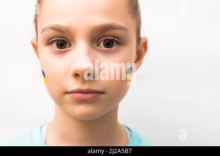 Gesicht eines verängstigten Mädchens, ein bemaltes Herz auf der Wange in gelb-blauen Farben der ukrainischen Flagge. Russlands Invasion der Ukraine, eine Bitte um Hilfe Stockfoto