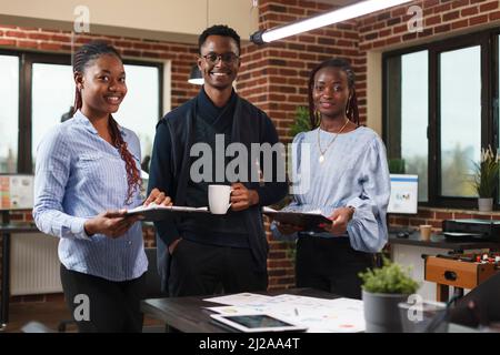 Professionelle Kollegen der Marketingagentur, die neben dem Tisch stehen, lächeln und die Kamera betrachten. Happy african american Business Company Team am Schreibtisch im modernen Bürobereich. Stockfoto