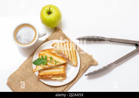 Hausgemachtes gegrilltes Käsesandwich zum Frühstück auf einem Teller mit einem Sacktuch, einer Tasse Kaffee, einer Küchenzange und einem Apfel. Draufsicht. Stockfoto