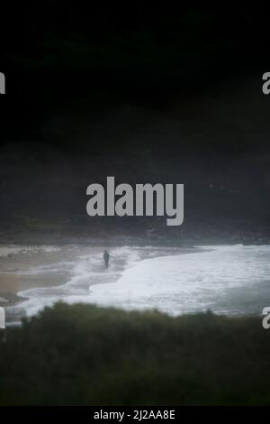 Blick auf die Küste durch ein Autofenster auf einen wild stürmischen grauen und feuchten Tag draußen mit einer winzigen Silhouette, die am Strand spazieren geht. Stockfoto